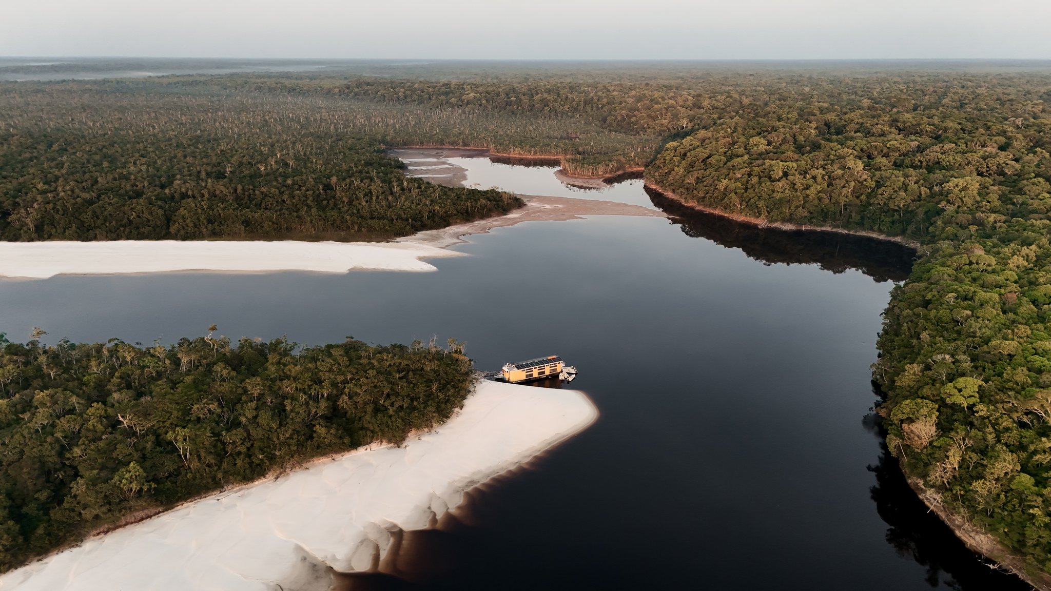 Aerial view of a solar-powered yellow houseboat moored on a broad white river sandbar, surrounded by dark blackwater channels and dense Amazon rainforest canopy, on a luxury Amazon cruise in Brazil