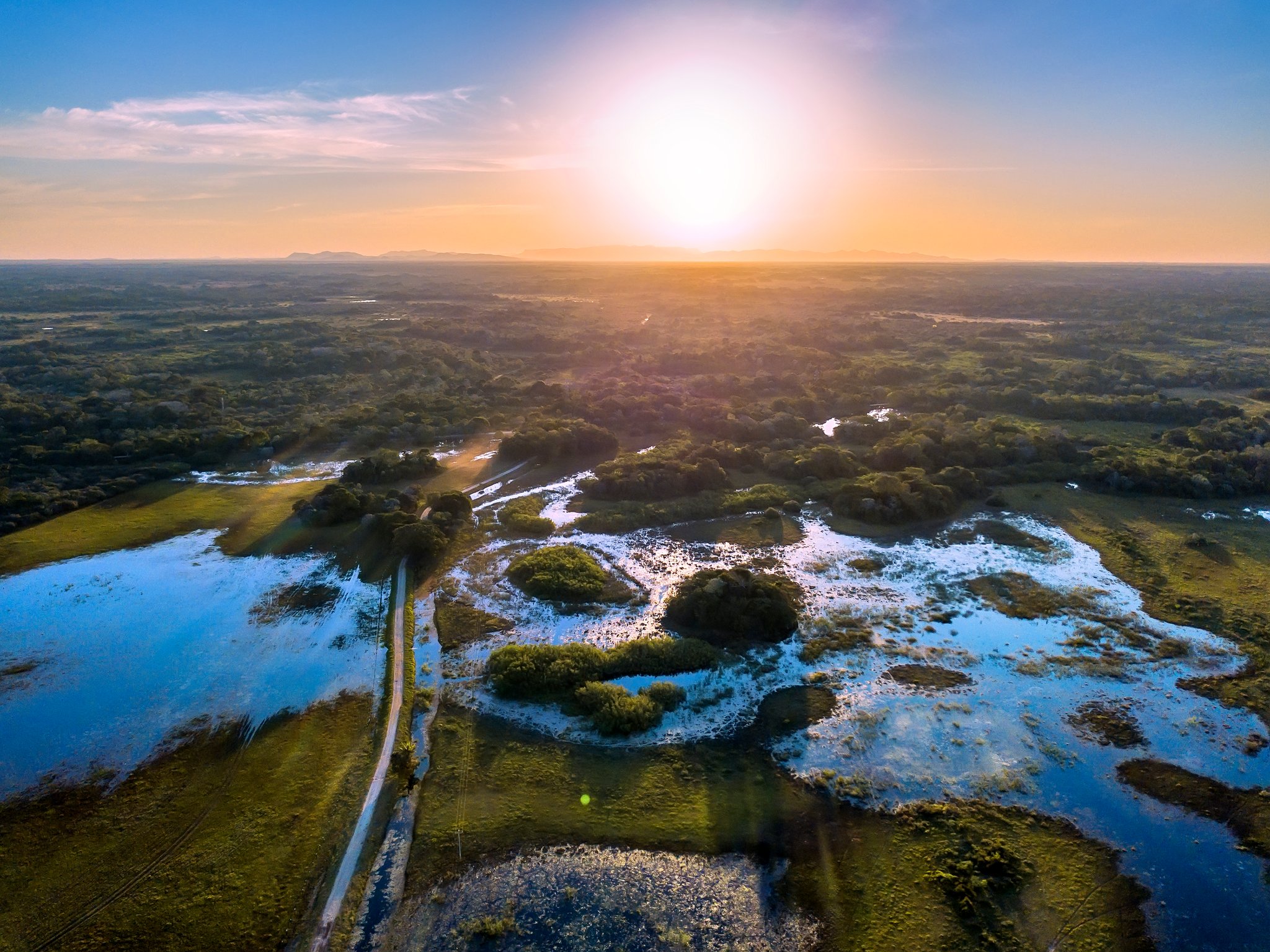 Aerial view of a southern pantanal safari landscape at sunset, showing flooded plains, scattered tree islands, a dirt track, and distant hills bathed in golden light