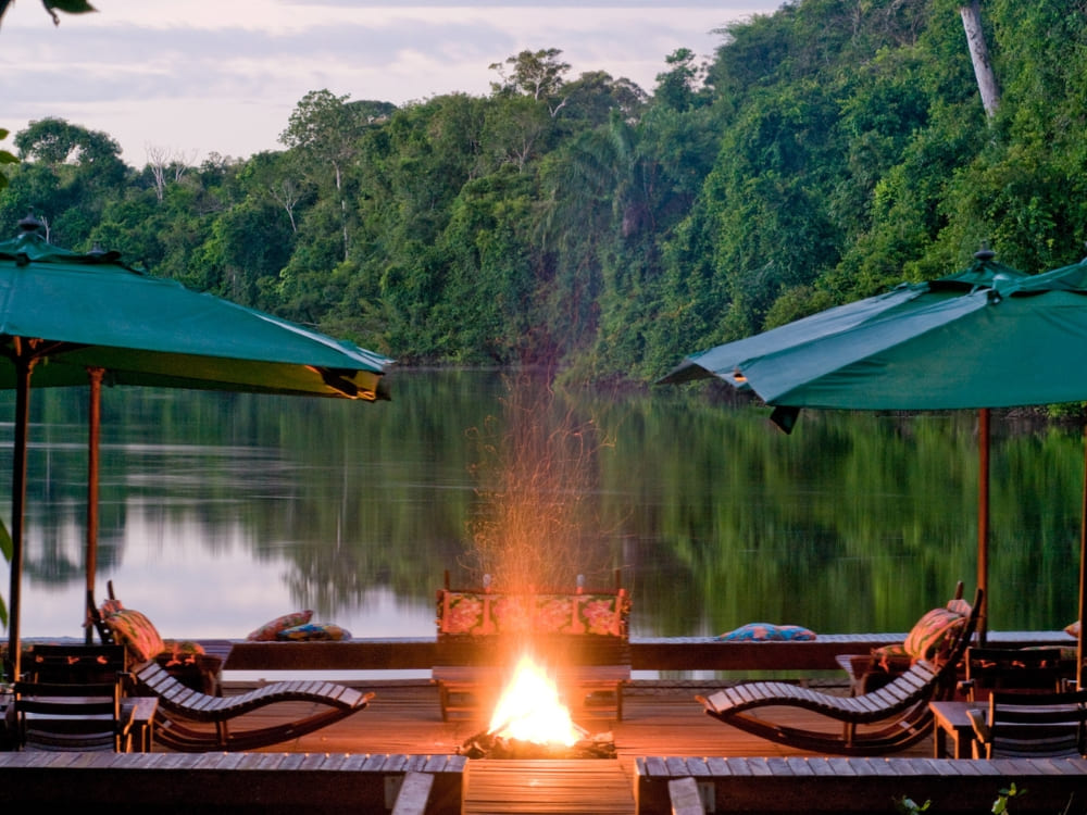 A riverside lodge deck at dusk with green umbrellas, sun loungers and a lit fire pit reflected in the calm Amazon river.