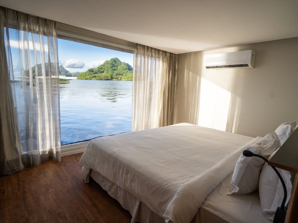 A bright lodge bedroom with a large bed and floor-to-ceiling windows overlooking a river and a forested island in the Amazon.