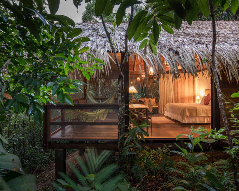 A luxury Amazon jungle lodge bungalow with a thatched roof and glass walls glowing with warm light at dusk, a hammock on its wooden deck amid the rainforest. A luxury Amazon jungle lodge bungalow with a thatched roof and glass walls glowing with warm light at dusk, a hammock on its wooden deck amid the rainforest.