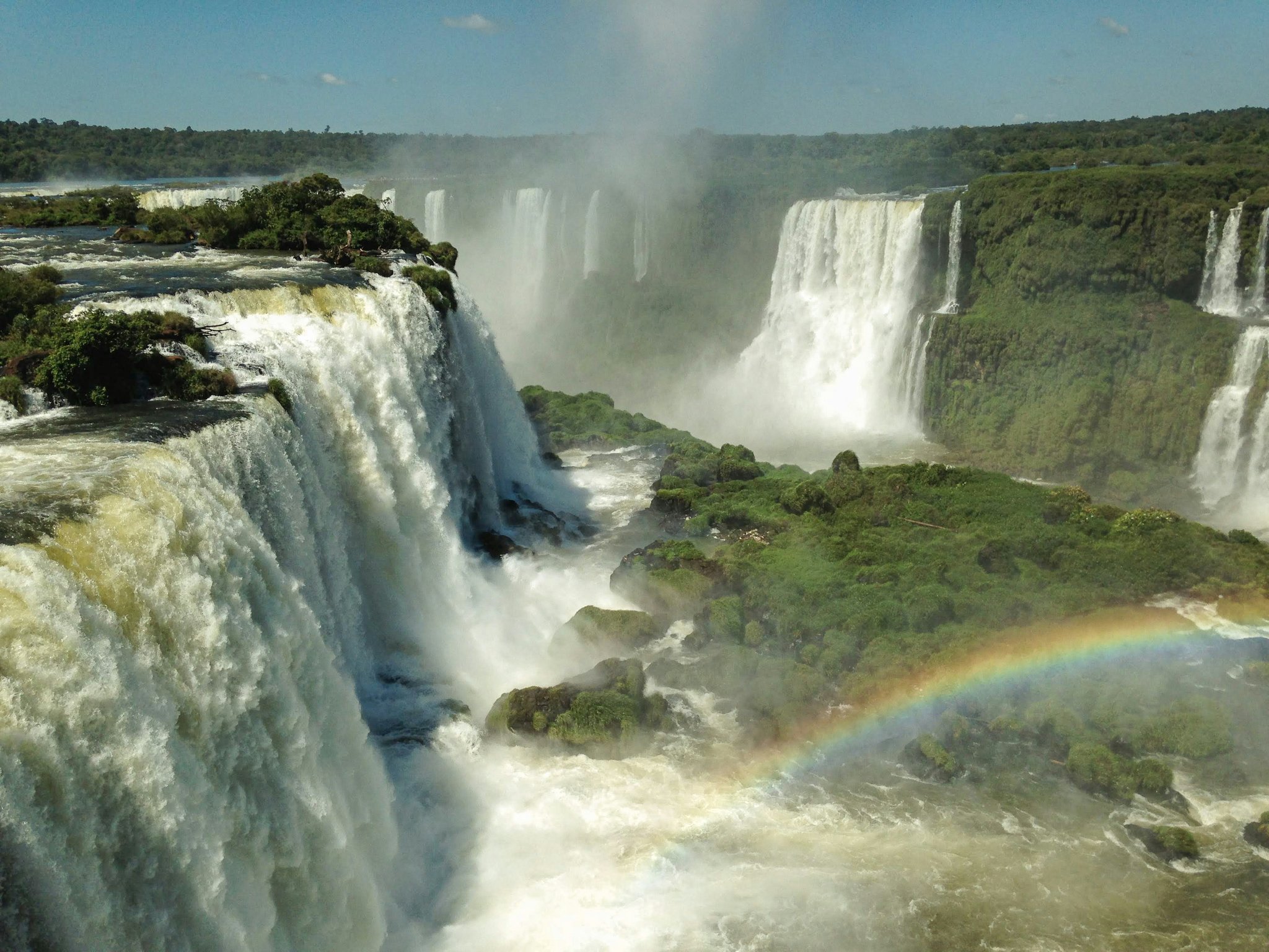 Panoramic view of Iguazu Falls on an iguazu falls private tour, with dozens of cascades plunging over lush green cliffs and a rainbow arching through the rising mist