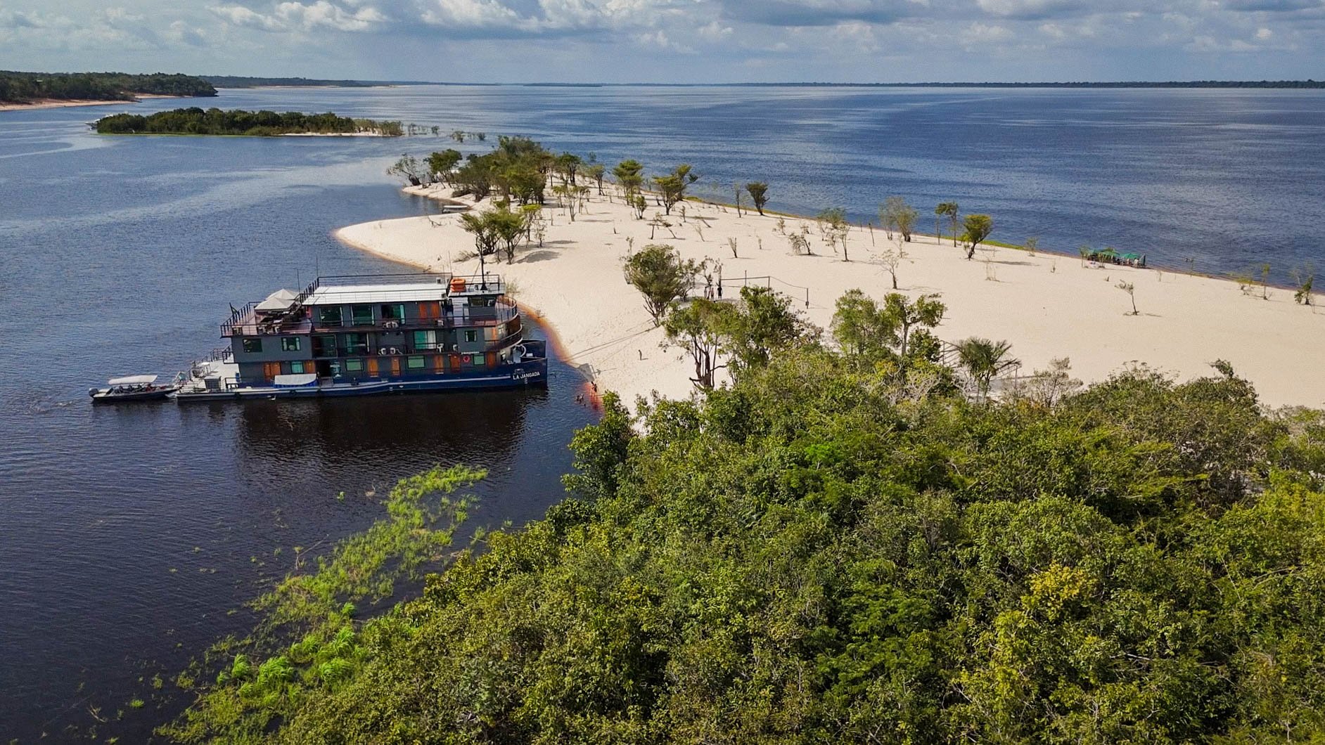 Aerial view of the La Jangada amazon cruise vessel moored beside a white sand beach on a dark-water river, surrounded by dense rainforest canopy and a wide river confluence under a partly cloudy sky.