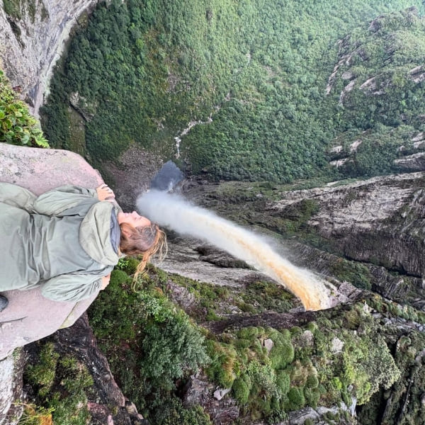 Eine Wanderin liegt am Felsrand über der Cachoeira da Fumaça und blickt in die Tiefe, während sich das Wasser über dem grünen Tal in Nebel auflöst, Chapada Diamantina, Brasilien.