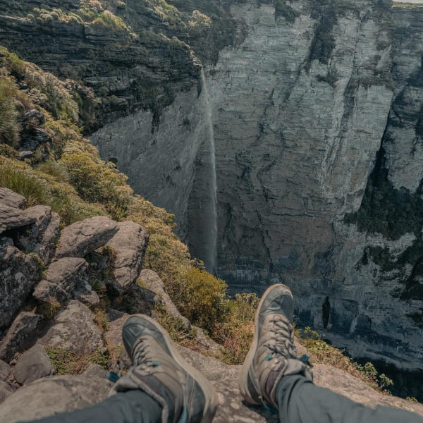Wanderschuhe an der Felskante am Aussichtspunkt der Cachoeira da Fumaça, während der Wasserfall gegenüber die Canyonwand hinabstürzt, Chapada Diamantina, Brasilien.