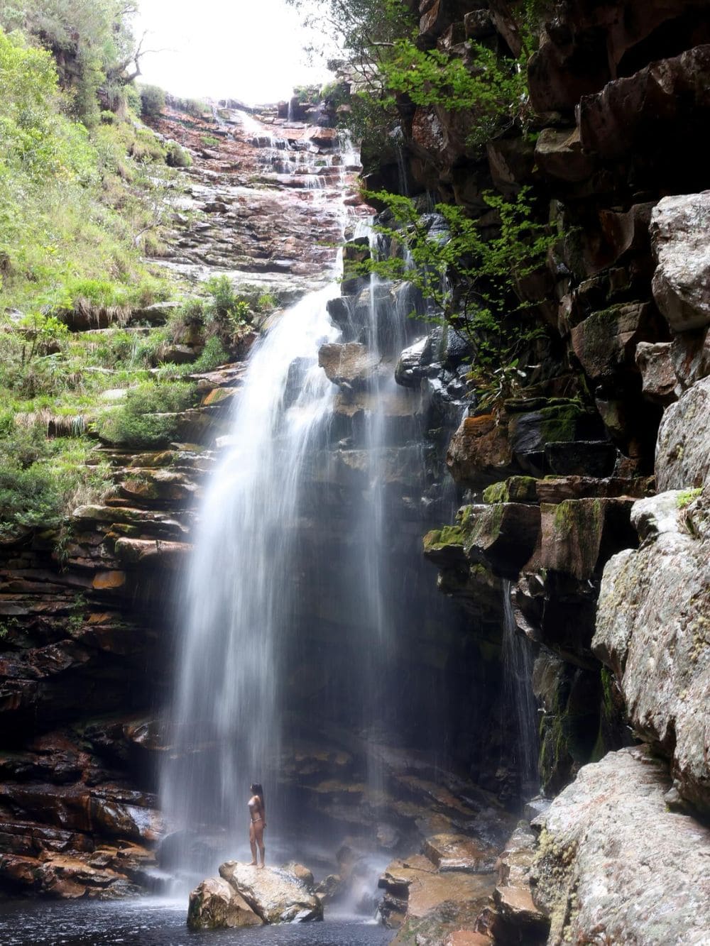 Eine Person steht auf einem Felsen am Fuß eines hohen Wasserfalls im Vale do Pati, Chapada Diamantina, Brasilien.