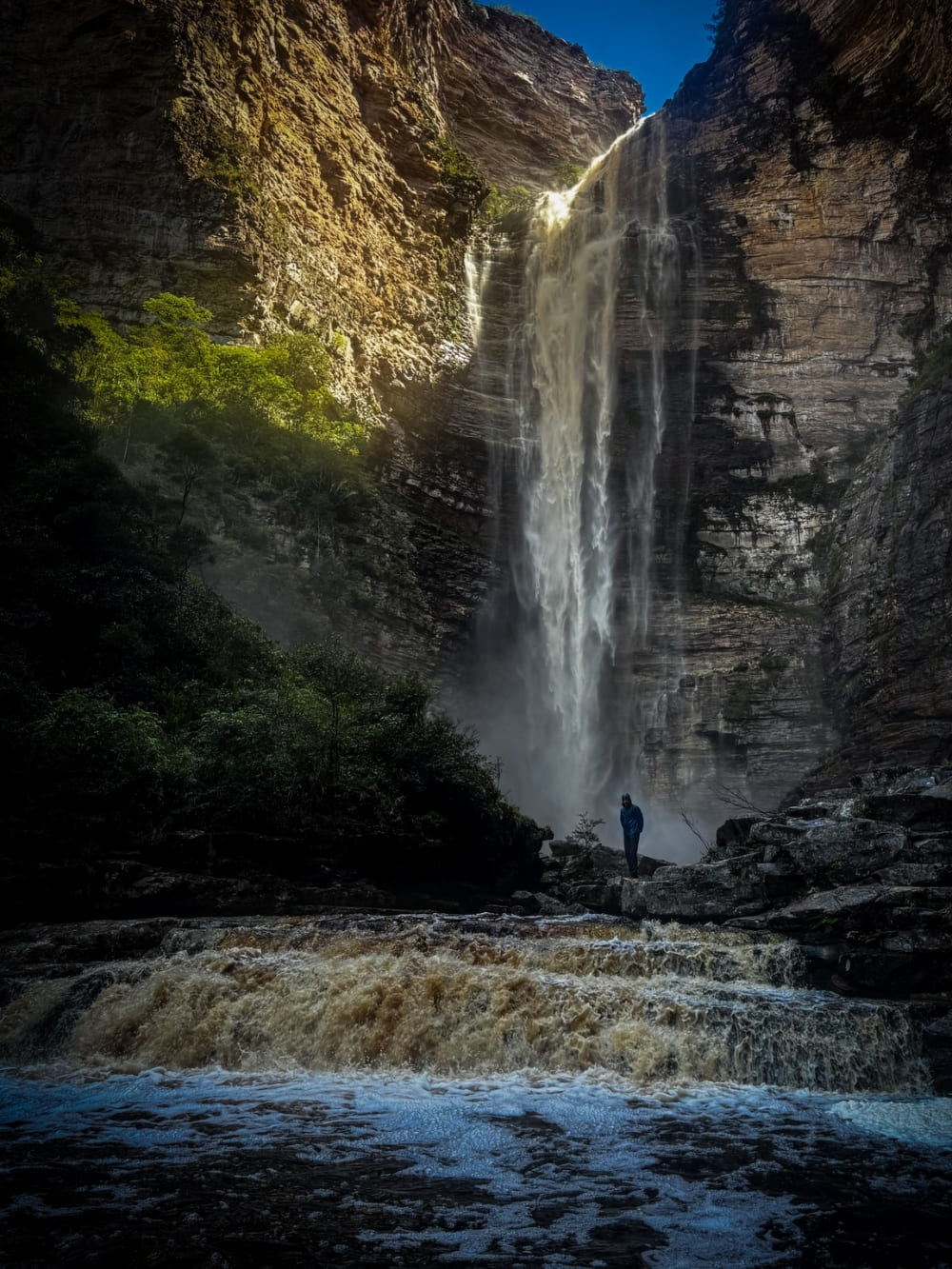 Ein einzelner Wanderer am Fuß der Cachoeira da Encantada, die zwischen den Canyonwänden über den bernsteinfarbenen Rio Samina stürzt, Chapada Diamantina, Brasilien.