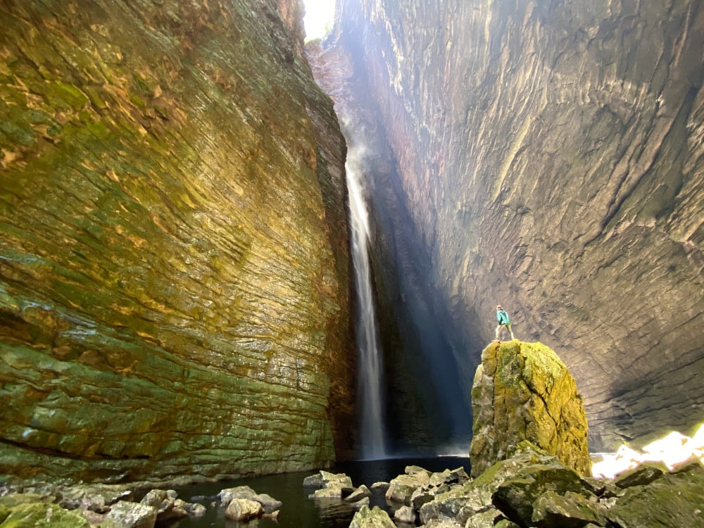 Ein Wanderer steht auf einem Felsblock vor dem Wasserfall Fumacinha in einem moosbewachsenen Canyon in der Chapada Diamantina, Brasilien.