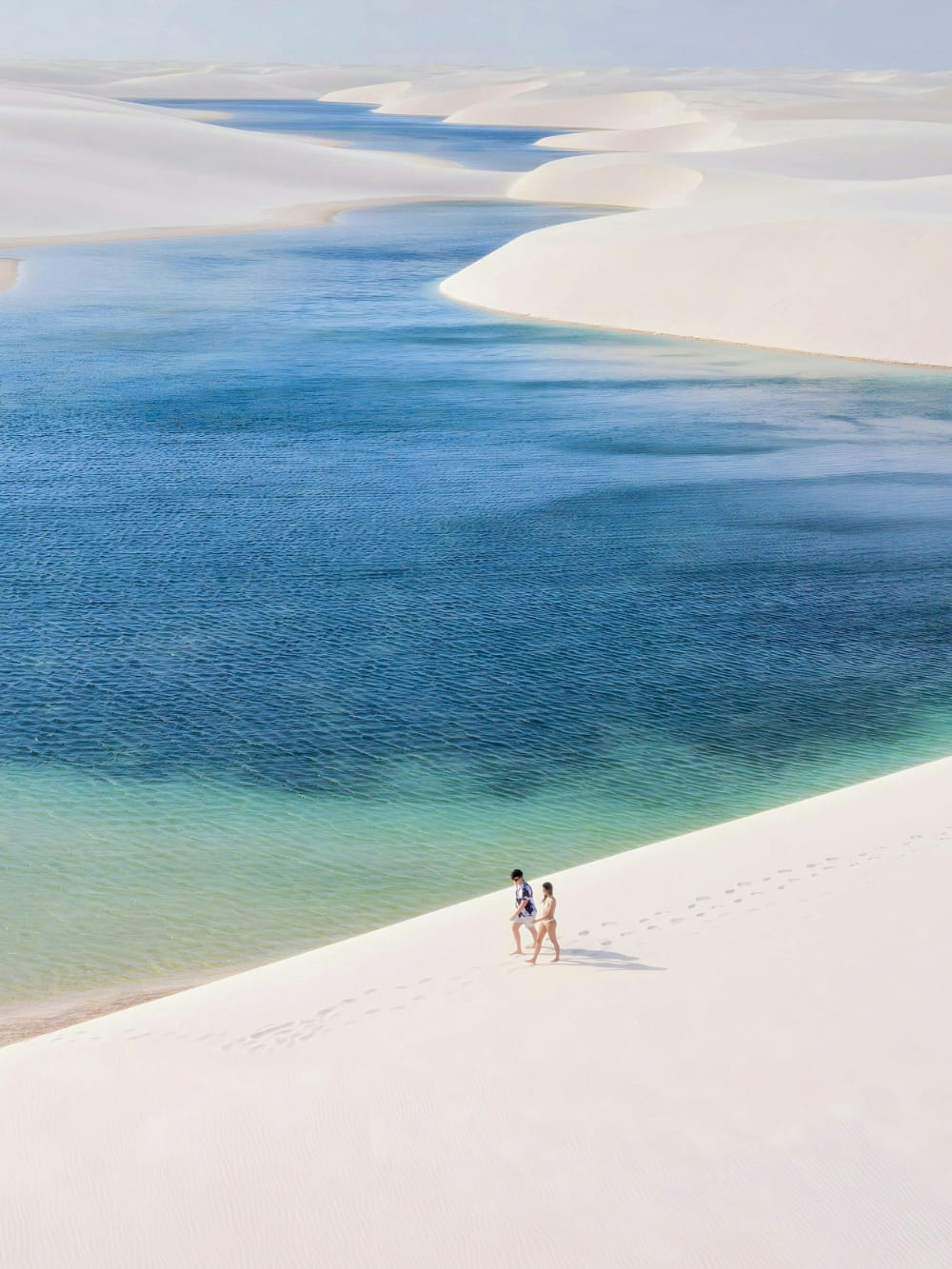 Ein Paar geht über eine weiße Sanddüne neben einer türkisfarbenen Lagune in den Lençóis Maranhenses, Brasilien.