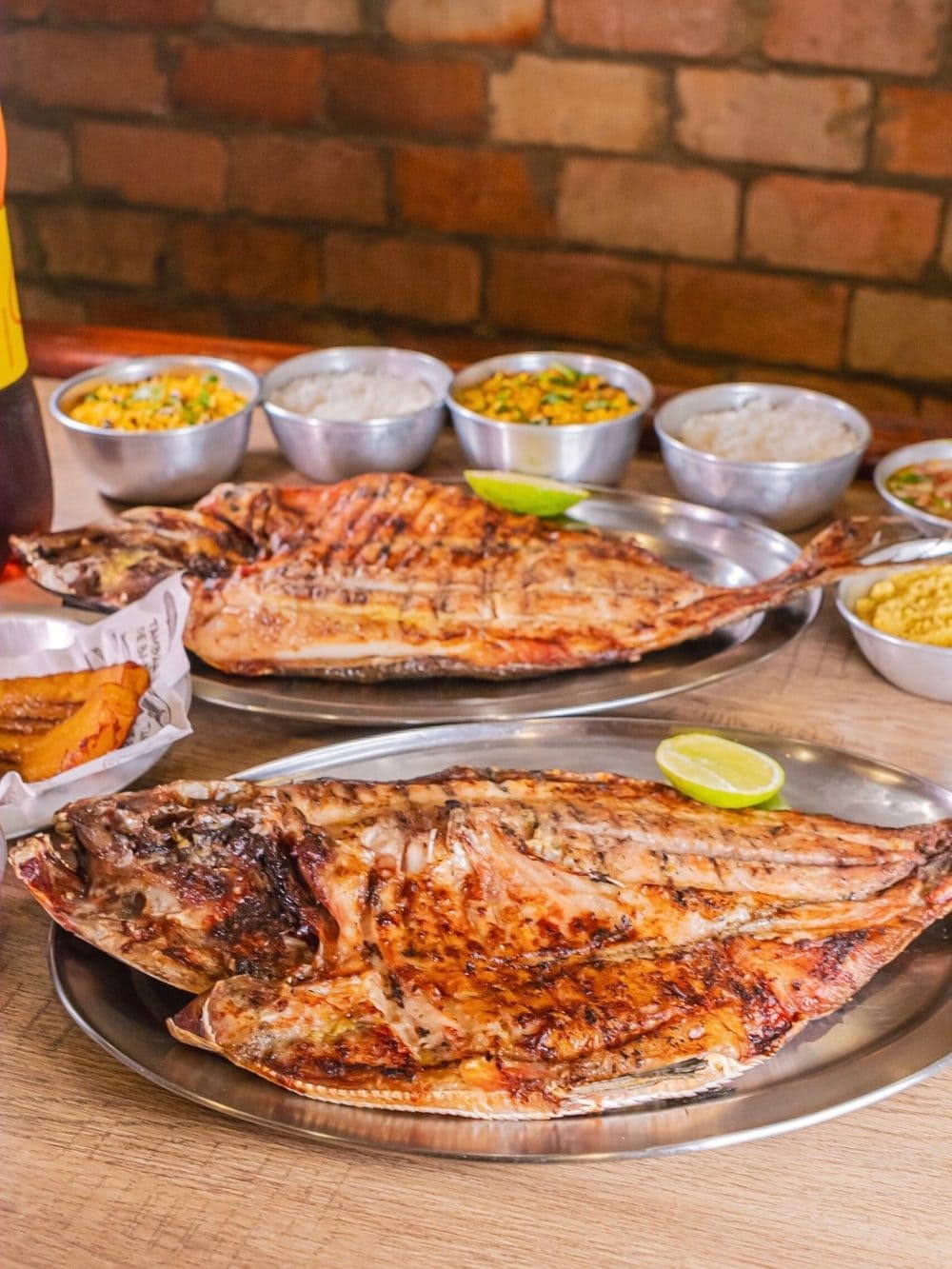 Zwei aufgeklappte gegrillte Tambaqui-Fische auf Metallplatten, dazu Limette, Reis und Farofa