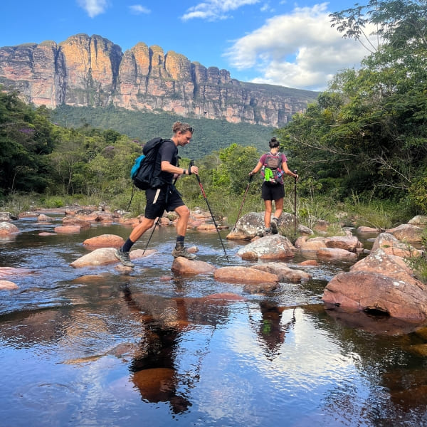 Zwei Wanderer durchqueren einen flachen Fluss unterhalb der Felswände der Chapada Diamantina, Brasilien.