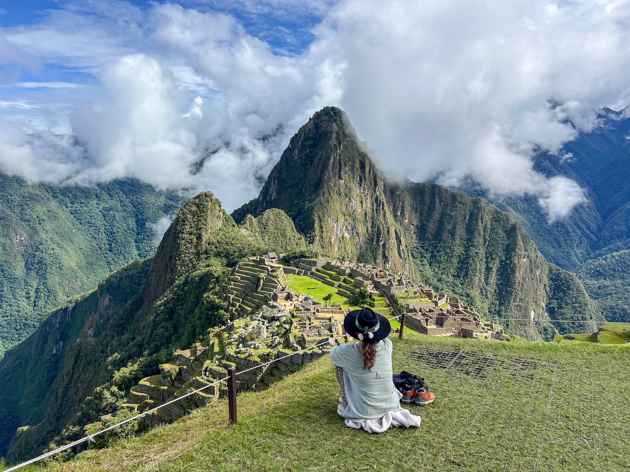 A woman in a wide-brimmed hat sits on a grassy ridge overlooking the Machu Picchu citadel during an Andes mountains tour, with Huayna Picchu peak and shifting clouds filling the valley behind her