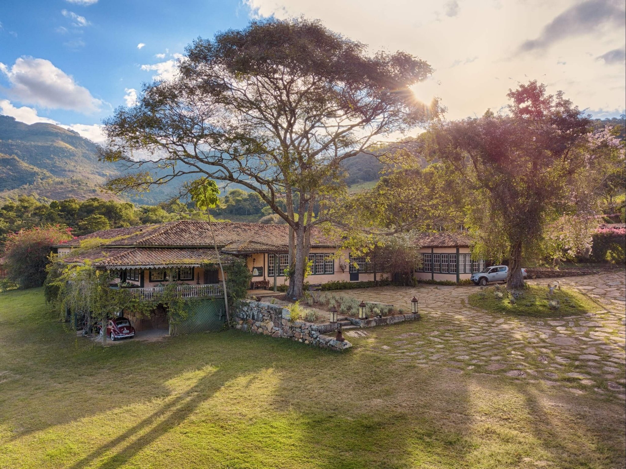 Aerial view of the Ibiti Project's Engenho Lodge, a colonial-style farmhouse with terracotta roof tiles, a wide veranda, and a cobblestone courtyard, framed by a large spreading tree and green mountain ridges in the background
