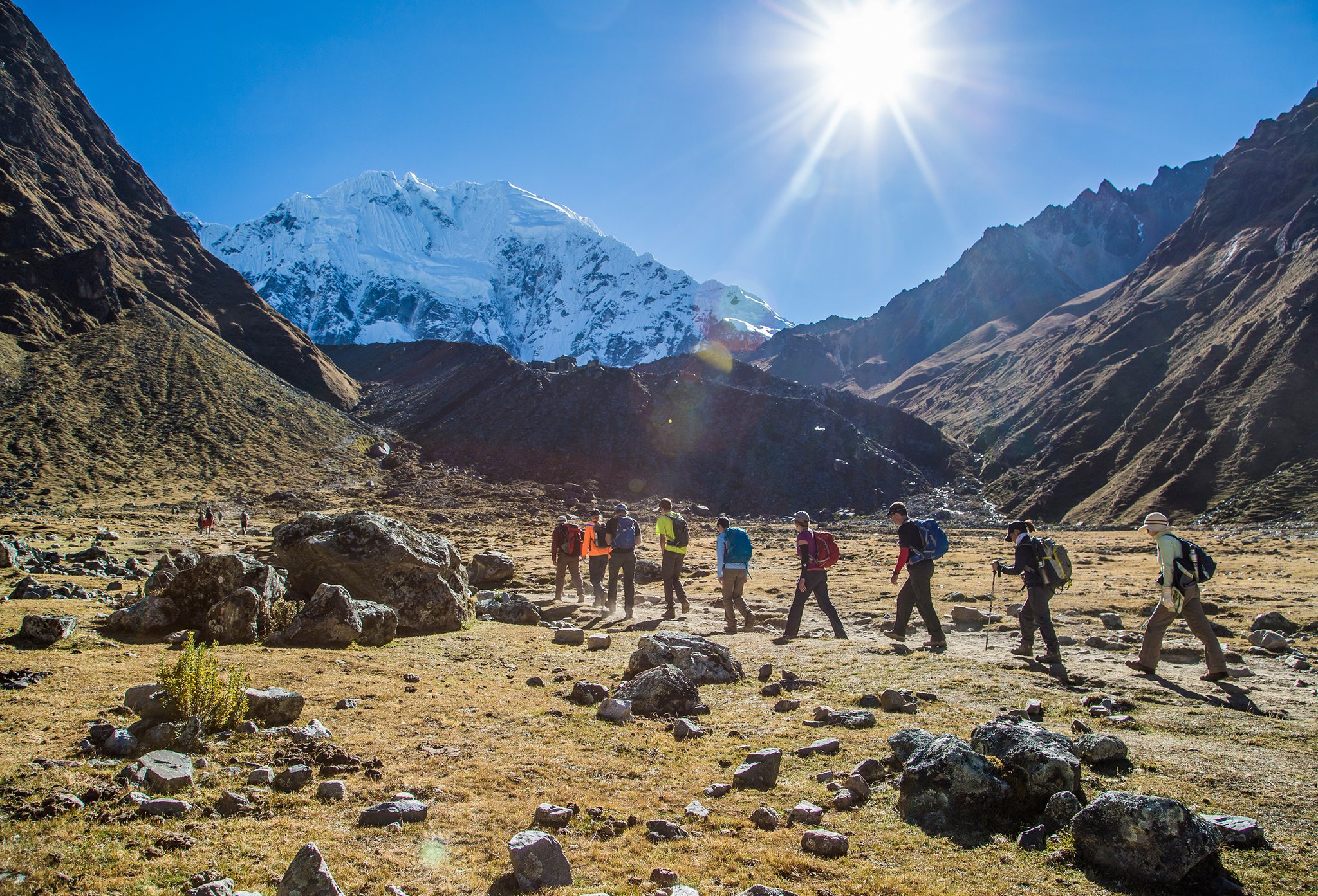 A group of trekkers with backpacks crosses a rocky high-altitude valley on the luxury Salkantay trek, with a snow-capped Andean peak rising under a bright sun in the background