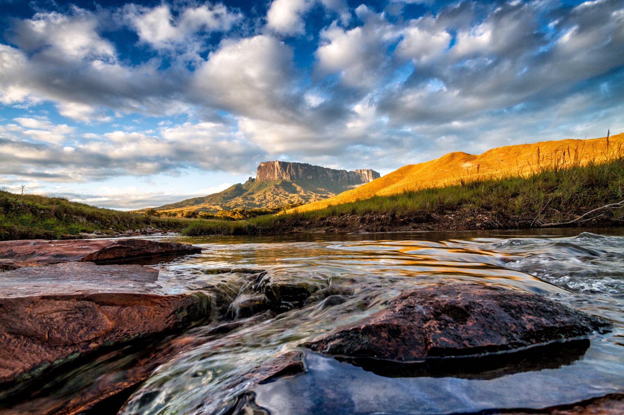 A shallow rocky river flows toward a flat-topped tepui on a mount roraima expedition, with golden savanna hills and a wide cloudy sky above