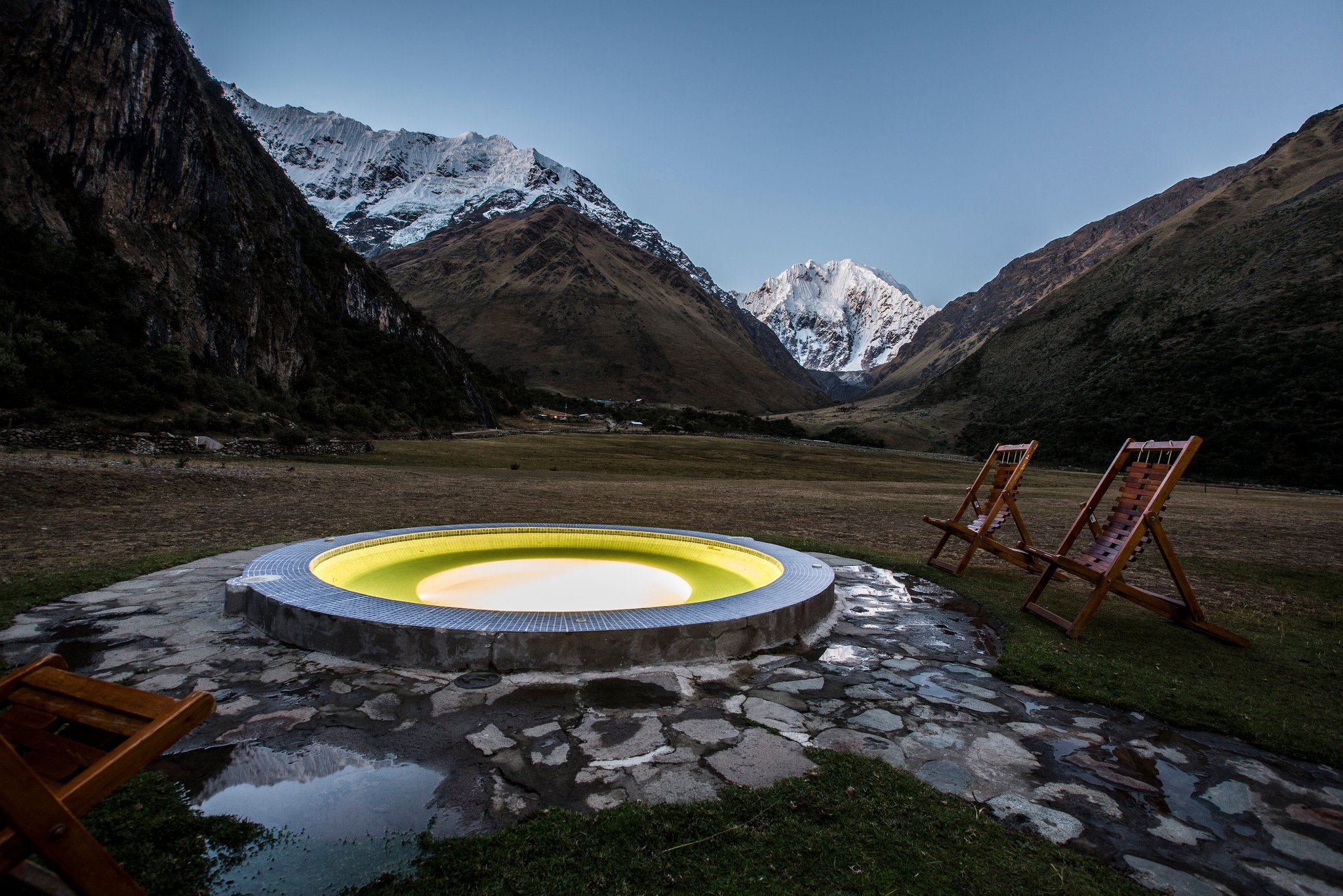 Illuminated circular outdoor hot tub at dusk on a stone terrace, part of the salkantay tour lodge experience, with wooden deck chairs and snow-capped Andean peaks rising across a wide mountain valley
