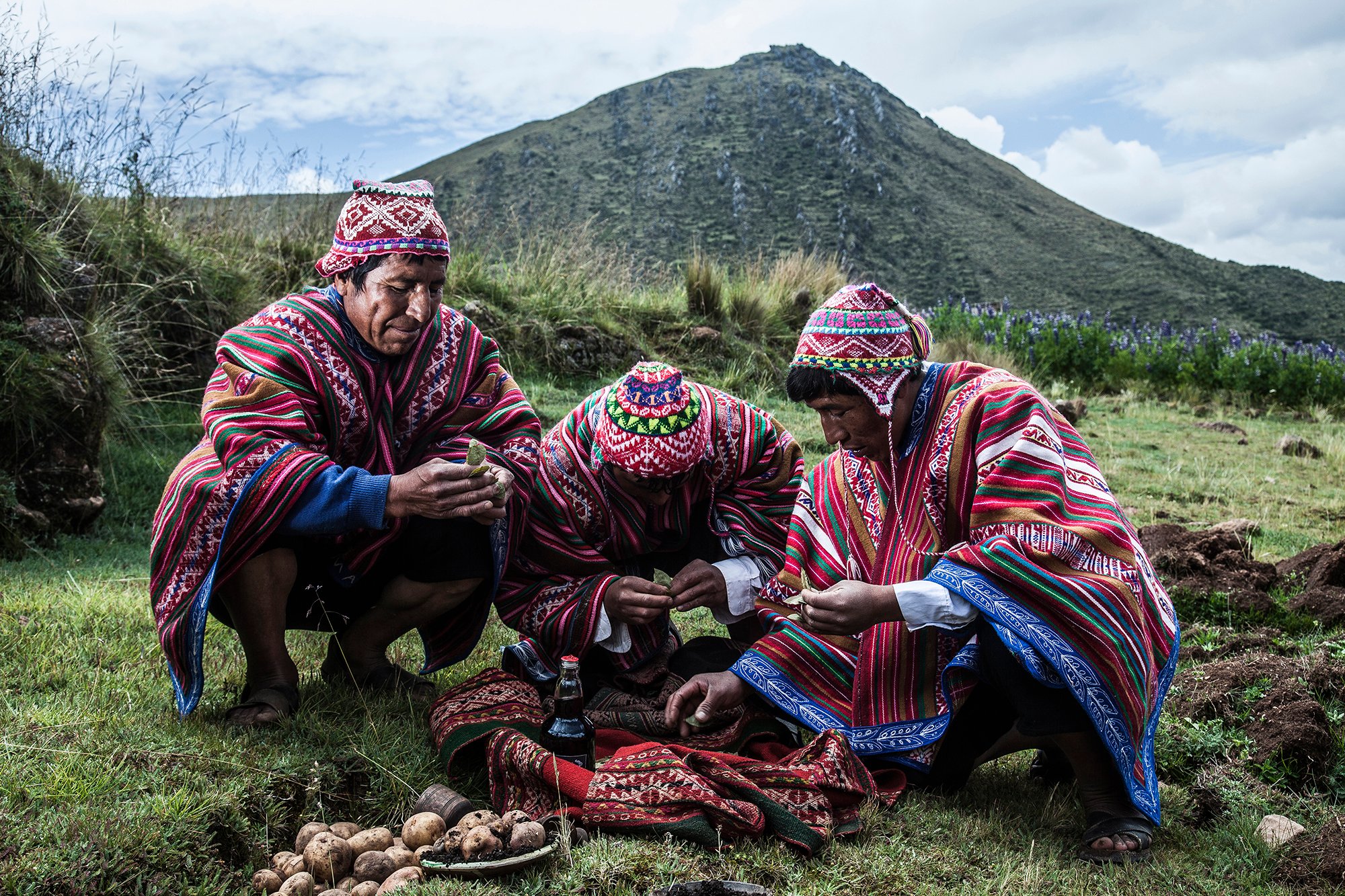 Three Andean men in traditional woven ponchos and chullos performing a ritual on a grassy hillside during a luxury Machu Picchu tour, with coca leaves, potatoes, and offerings spread on a cloth before them and a steep mountain peak rising behind