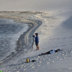 Cameraman filming from a tripod on a dune beside a lagoon while two people lie on the sand