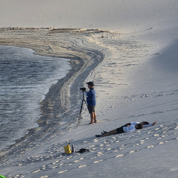 Cameraman filming from a tripod on a dune beside a lagoon while two people lie on the sand