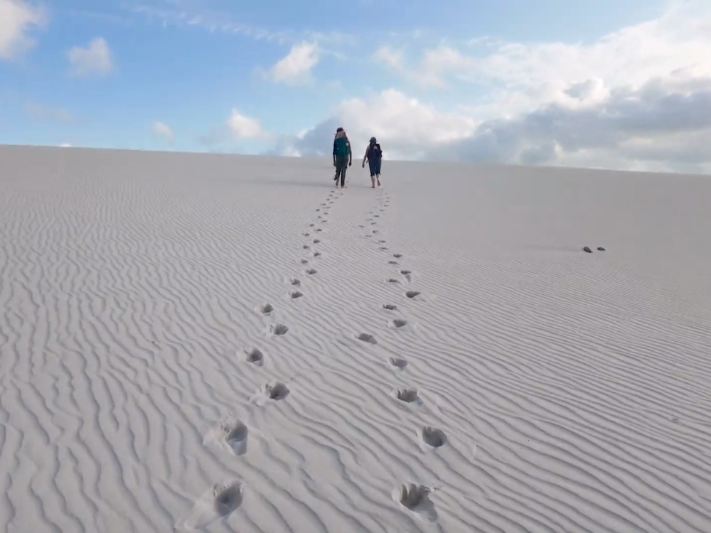 Dos excursionistas cruzan una duna blanca ondulada en los Lençóis Maranhenses, dejando un rastro de huellas