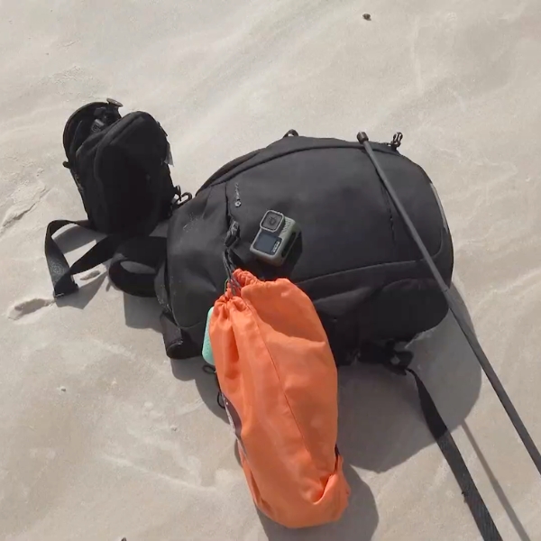 Mochilas, una cámara de acción y una bolsa estanca naranja sobre la arena durante una parada del trekking