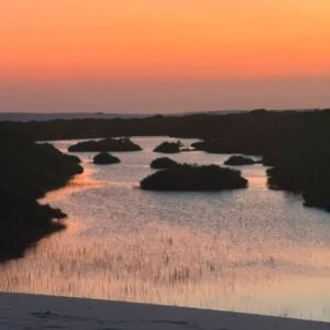Orange sky reflected on a lagoon dotted with islands of vegetation, seen from a dune in Lençóis Maranhenses