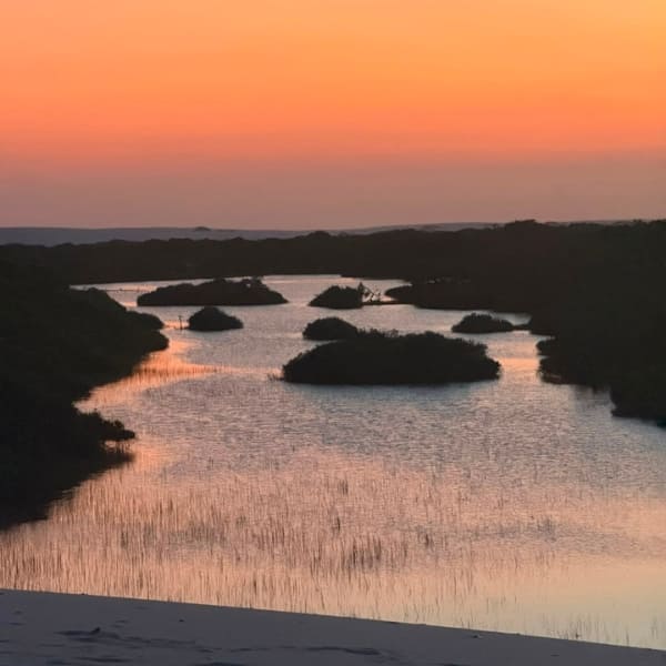 Orange sky reflected on a lagoon dotted with islands of vegetation, seen from a dune in Lençóis Maranhenses