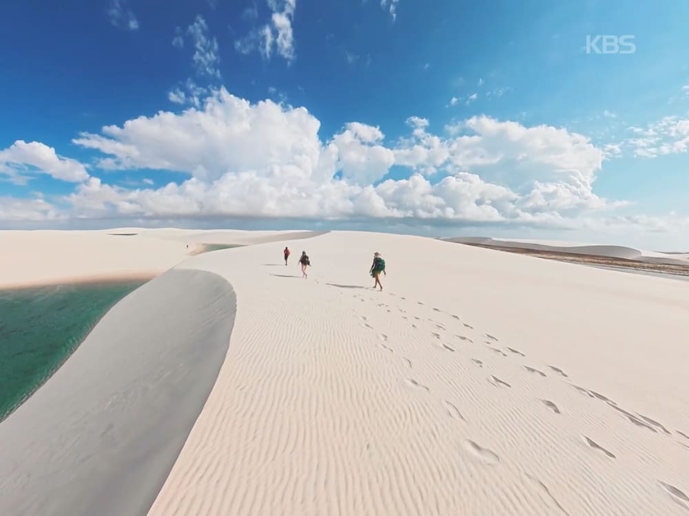 Trekkers walking along a white sand dune next to a turquoise lagoon in Lençóis Maranhenses