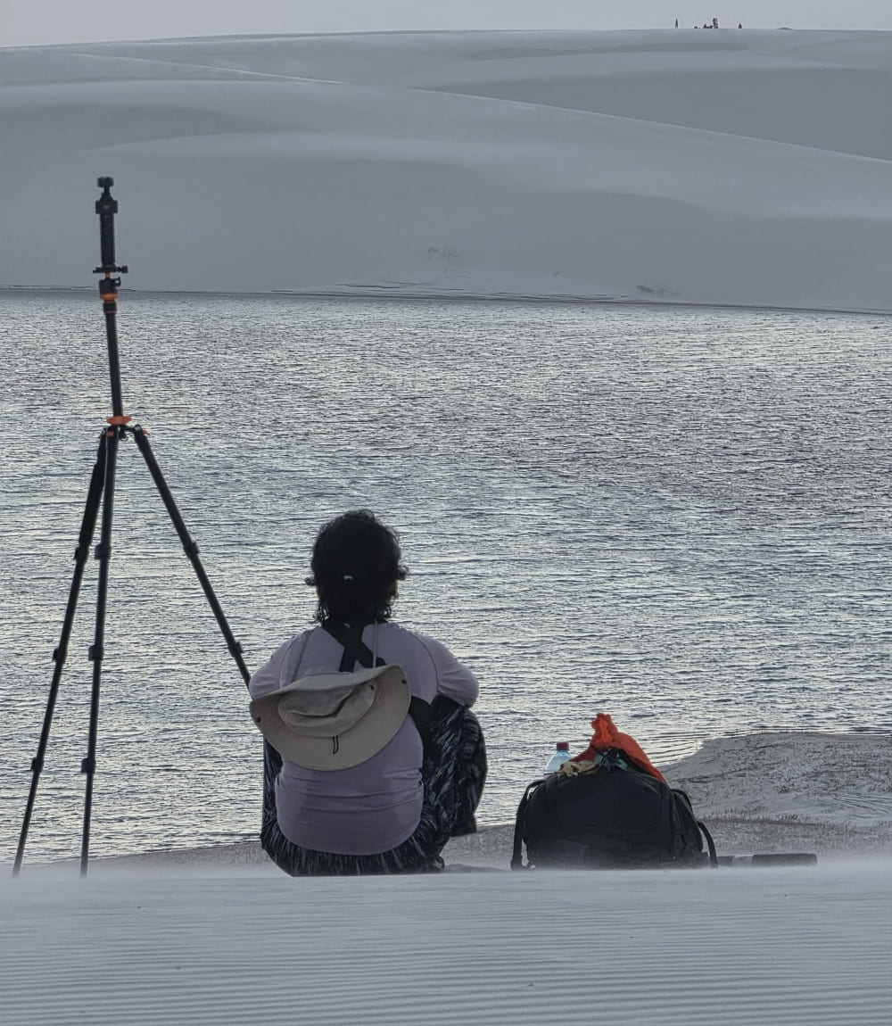 Person sitting on a dune beside a camera tripod, facing a lagoon in Lençóis Maranhenses