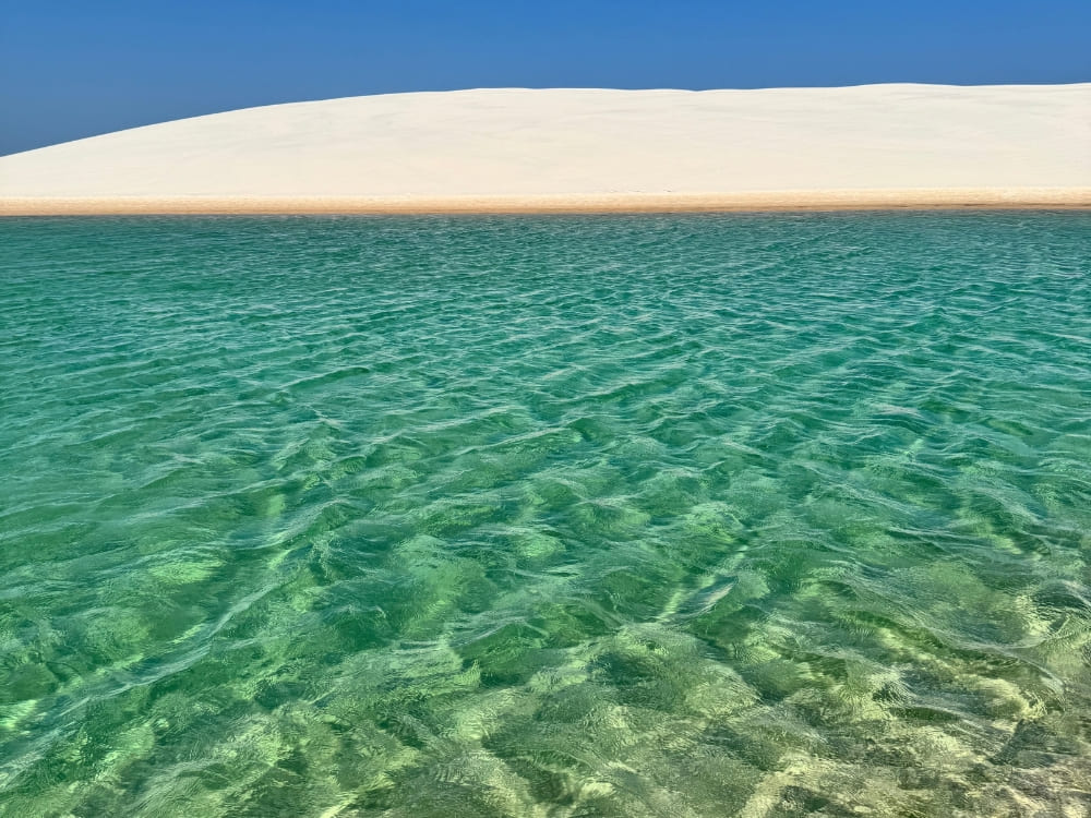 Eau verte et cristalline d’une lagune devant une dune de sable blanc sous un ciel bleu aux Lençóis Maranhenses