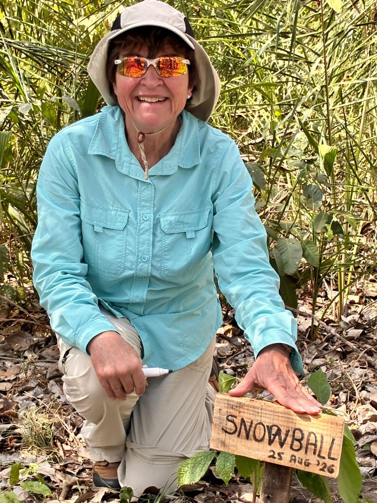 Andrea smiling beside the tree she planted at Caiman, marked with a sign reading Snowball, 25 Aug '26